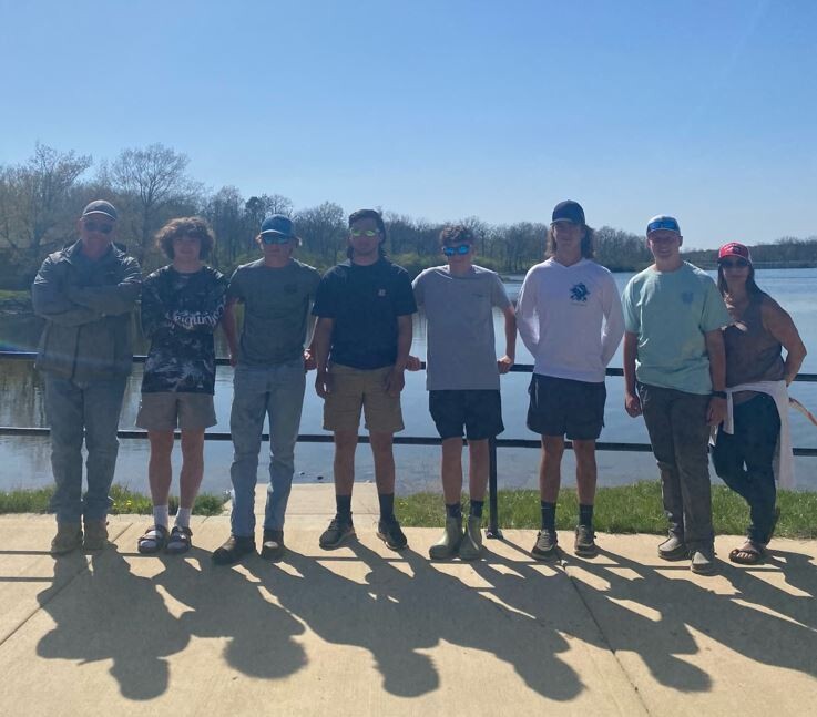 high school fishing team in caps poses in the sun by the lake