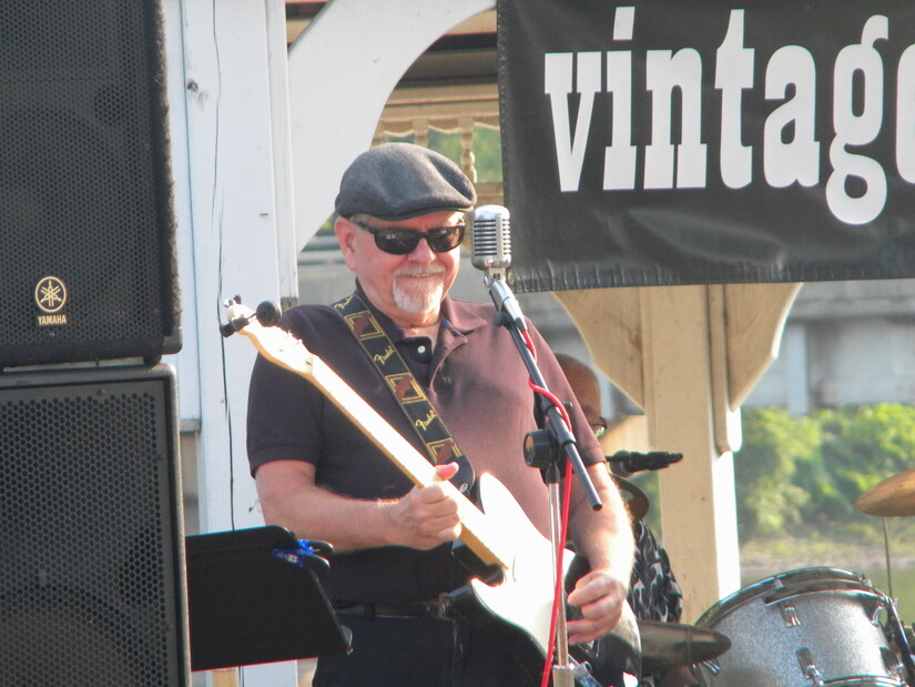 Guitar player with gray cap, sunglasses, and gray beard hits a guitar lick