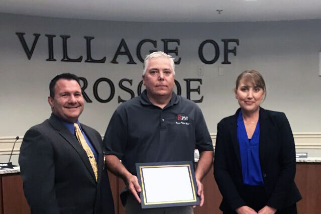 Man receives award plaque from the Village of Roscoe, flanked by two police officers