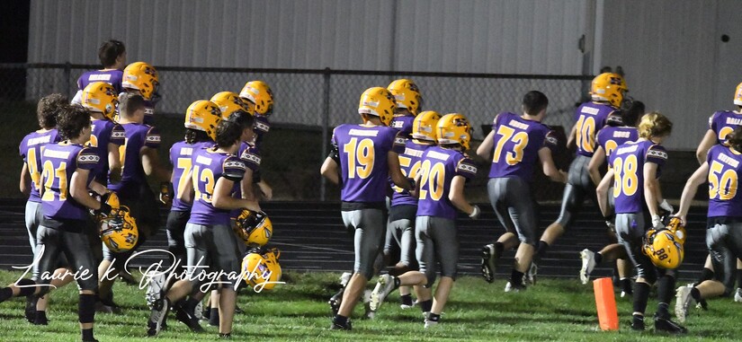 High school football team in purple and gold uniforms runs along the field.