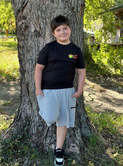 Boy in black shirt and shorts poses in front of tree trunk.