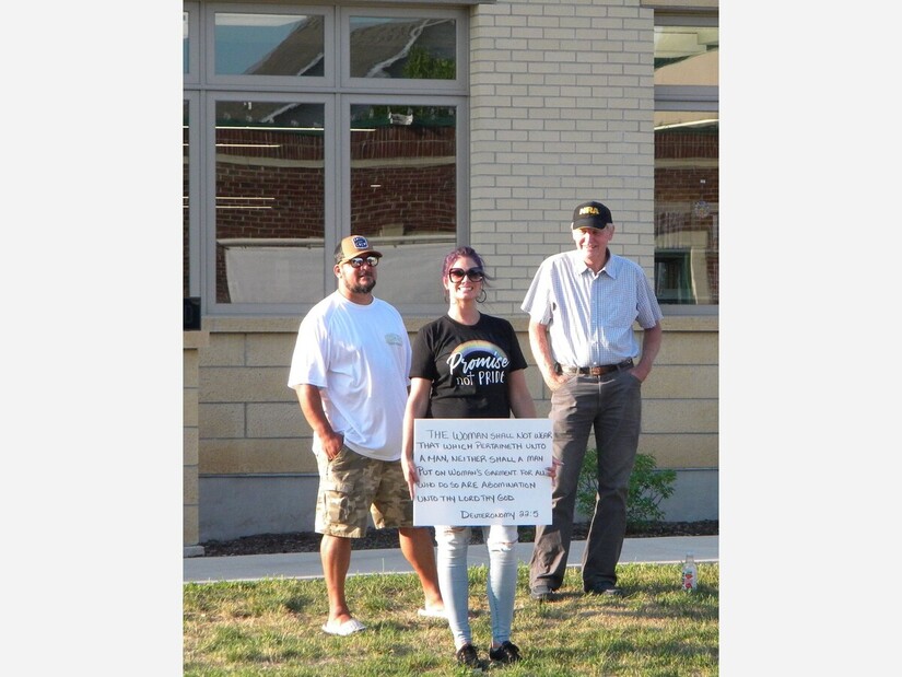 An anti-drag protester holds a sign quoting Deuteronomy 22:5, flanked by two men.