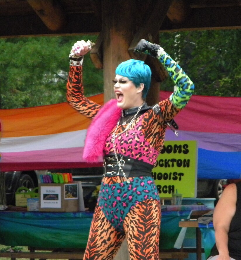 Drag queen with blue hair and tiger print clothes lip-synch at a park with her arms raised.